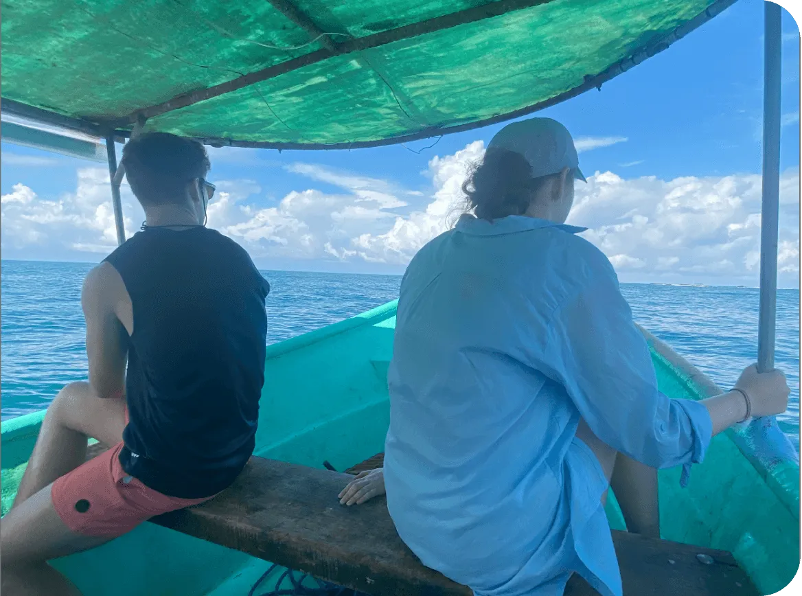 Two people sit on a wooden bench inside a small turquoise boat, looking out at calm ocean water beneath a bright blue sky and green canopy.