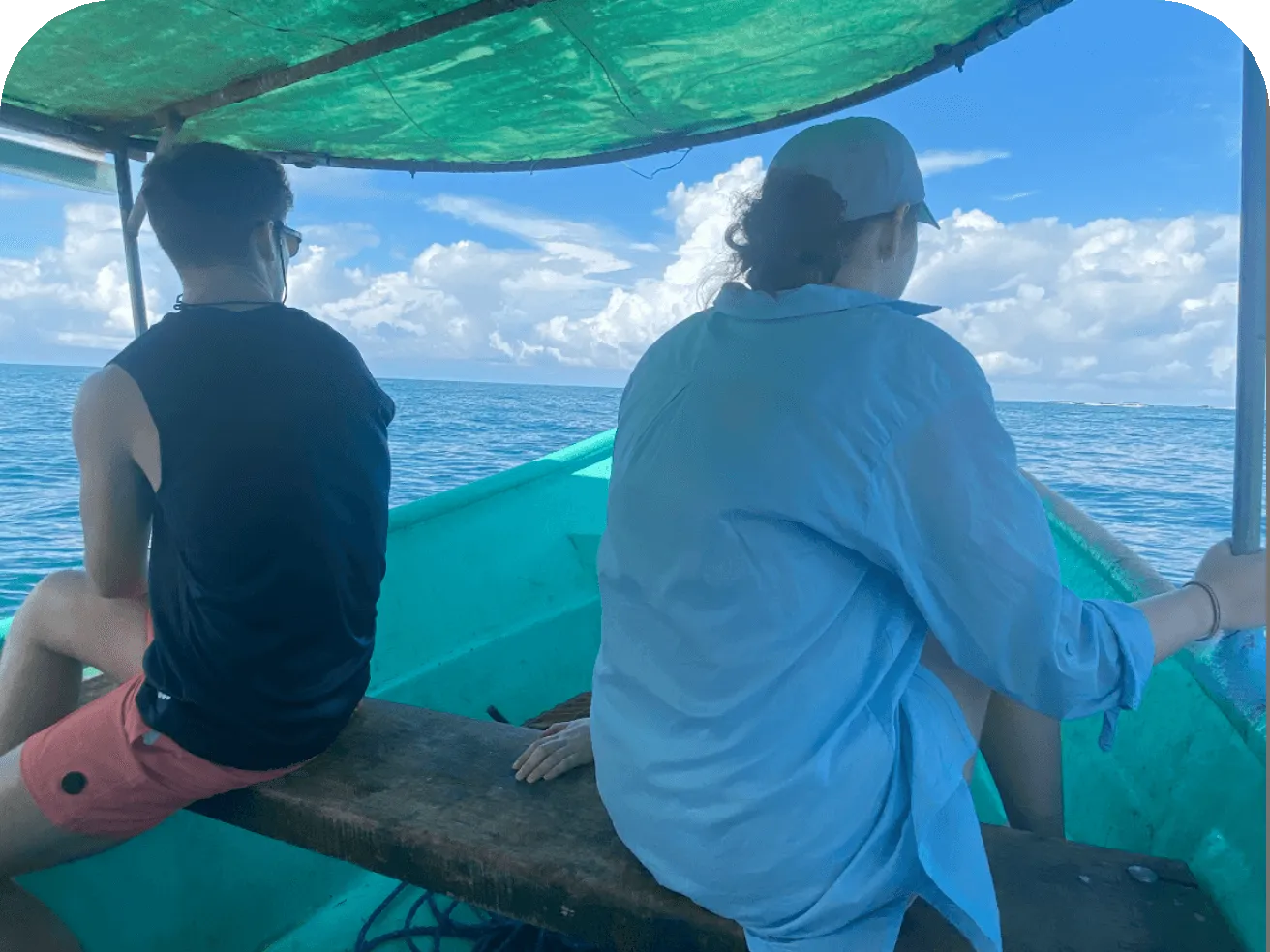 Two people sit on a wooden bench inside a small turquoise boat, looking out at calm ocean water beneath a bright blue sky and green canopy.