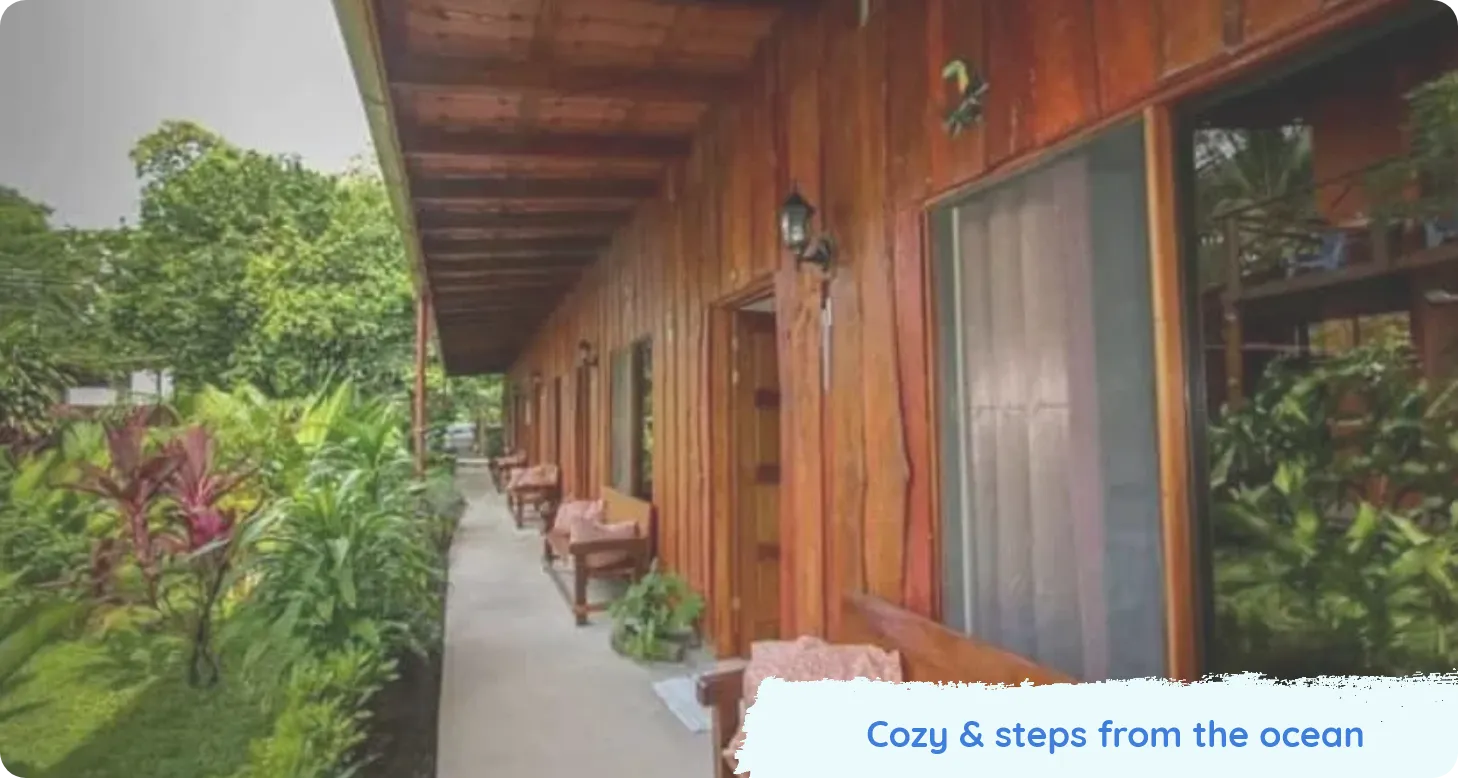 Exterior walkway of a wooden building surrounded by tropical plants. Text reads Cozy & steps from the ocean.